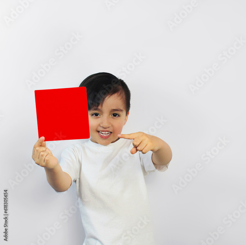 Child with big smile holds a red piece of paper points at it with his finger. in a white t-shirt on a white background, Space for text, banners, label surfaces, and ads. Red card, stop, signal