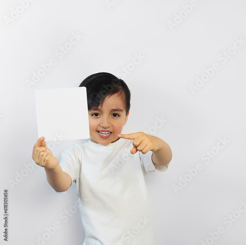 A latin child holds a white piece of paper points at it with his finger.  Wears a white t-shirt on a white background, Space for text, banners, label surfaces, and ads.