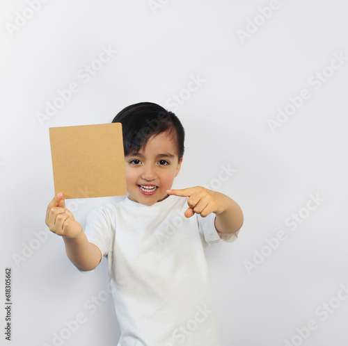 A kid with big smile holds a brown piece of paper in a white t-shirt on a white background, points at it with his finger. Space for text, banners, label surfaces, and ads. craft color