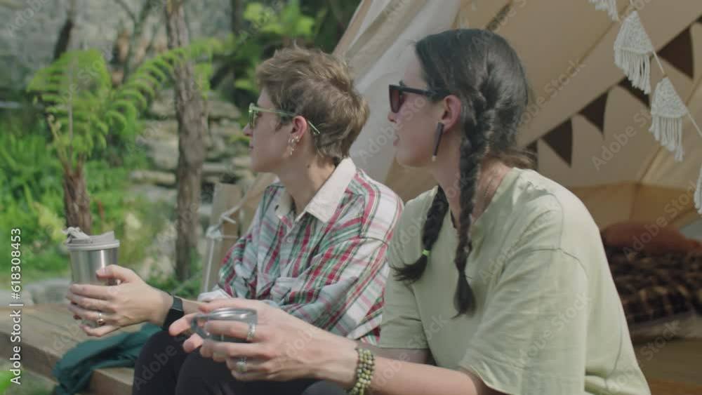 Medium shot of two female tourists in sunglasses sitting outside of glamping tent, holding tea mug and thermos and discussing nature around on summer vacation Medium shot of two female tourists in sunglasses sitting outside of glamping tent, holding tea mug and thermos and discussing nature around on summer vacation