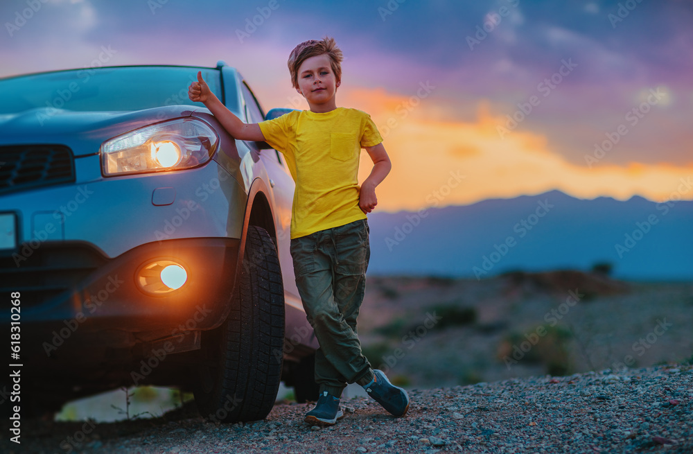 Naklejka premium Successful boy standing by the car and showing thumbs up at twilight on mountains background