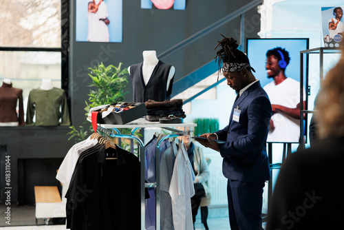 African american employee checking clothes inventory on tablet computer, looking at fashionable merchandise on hangers. Showroom worker checking discount list on device in shopping centre