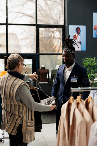 Senior customer discussing black bag material with worker, shopping for casual wear in modern boutique. Elderly client buying fashionable clothes and stylish accessories in clothing store