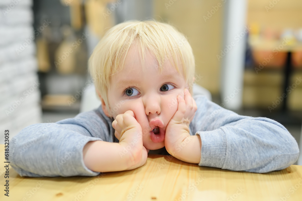 Funny emotional child is at a table in a cafe. Cute little boy sitting in restaurant and waiting for his order. Kid tired of waiting and is making faces. Sincere behavior of children in public places Funny emotional child is at a table in a cafe. Cute little boy sitting in restaurant and waiting for his order. Kid tired of waiting and is making faces. Sincere behavior of children in public places