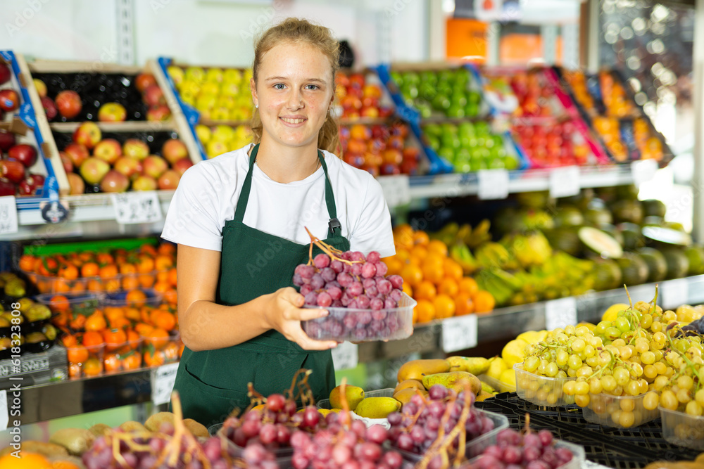 Positive young girl seller in uniform holding ripe red grape in grocery shop