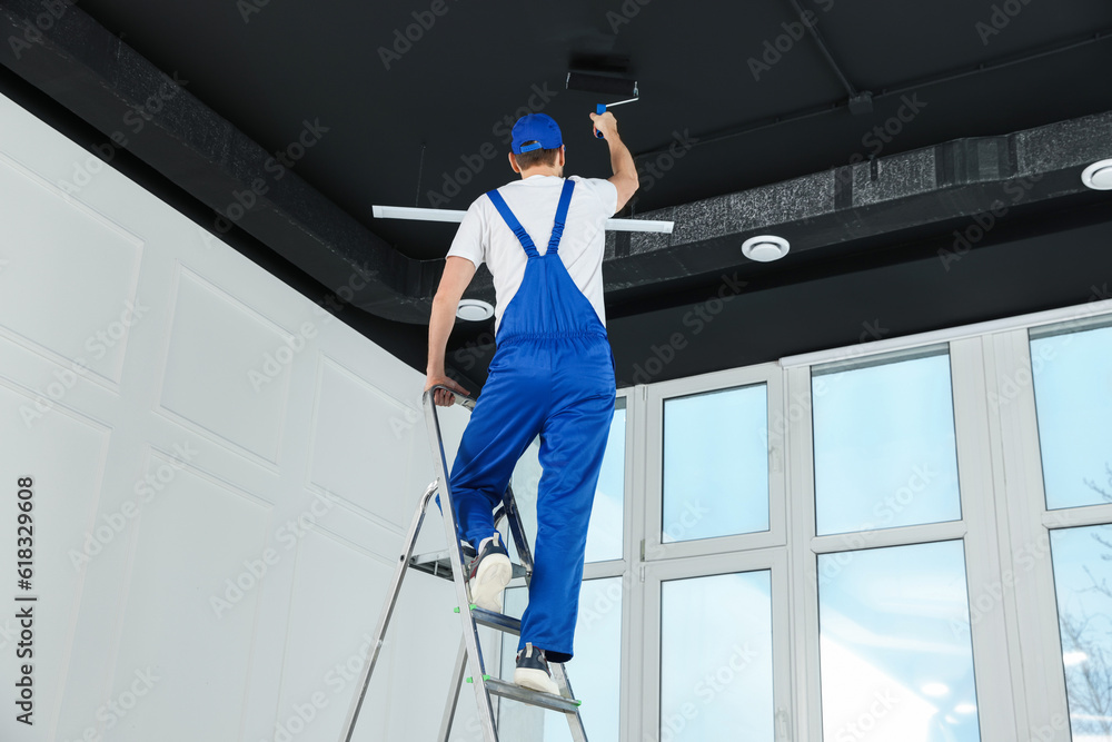 Worker in uniform painting ceiling with roller indoors, back view