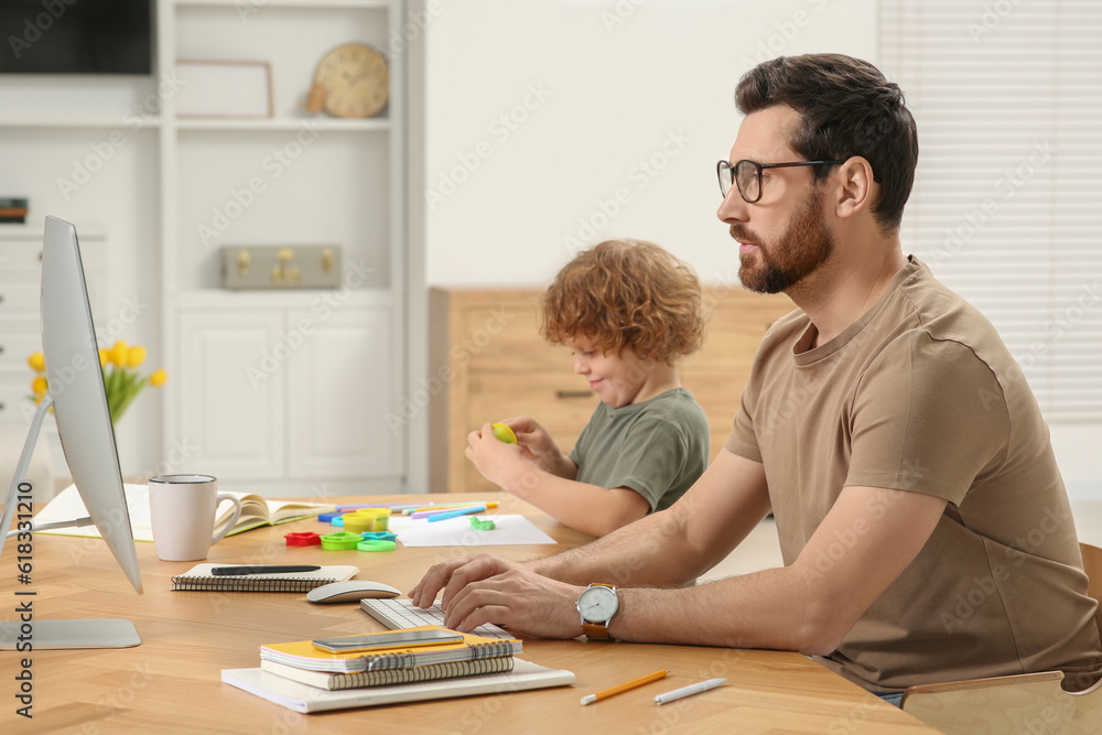 Man working remotely at home. Father using computer while his son playing at desk Man working remotely at home. Father using computer while his son playing at desk