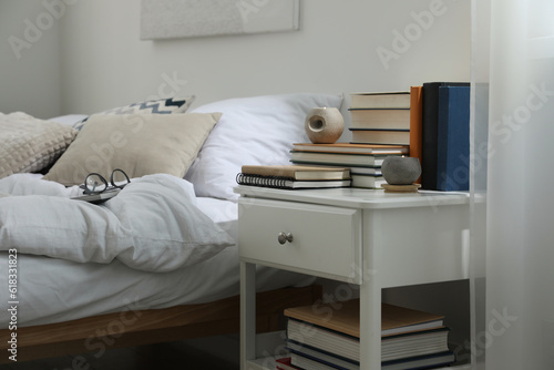 Hardcover books and scented candles on white bedside table in bedroom