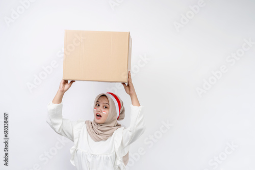 Cheerful Asian muslim woman celebrating indonesian independence day carrying package box isolated on white background. Indonesian independence day on 17 august concept