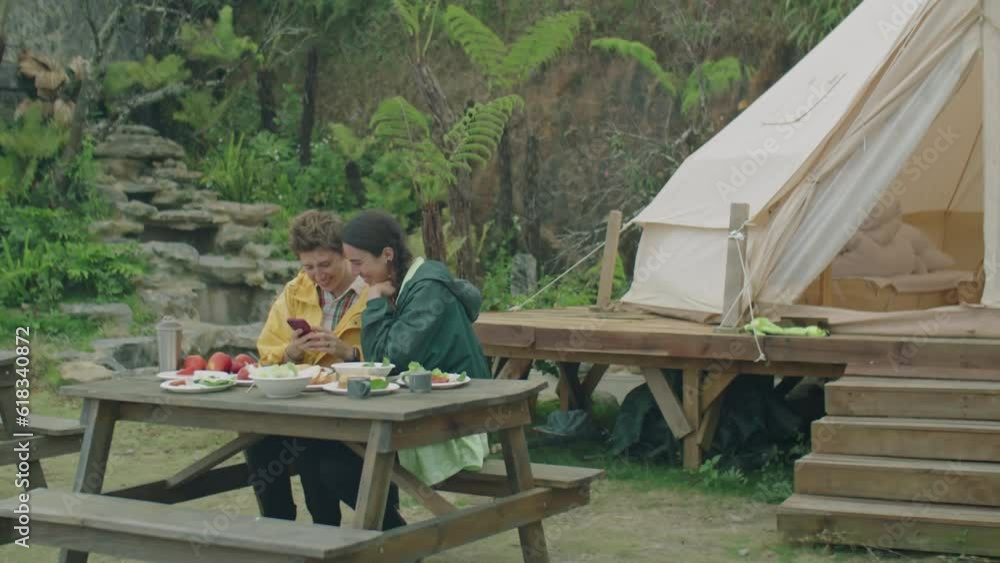 Full shot of two girlfriends sitting at table outdoors in front of glamping tent and taking selfie with smartphone while having dinner in nature Full shot of two girlfriends sitting at table outdoors in front of glamping tent and taking selfie with smartphone while having dinner in nature