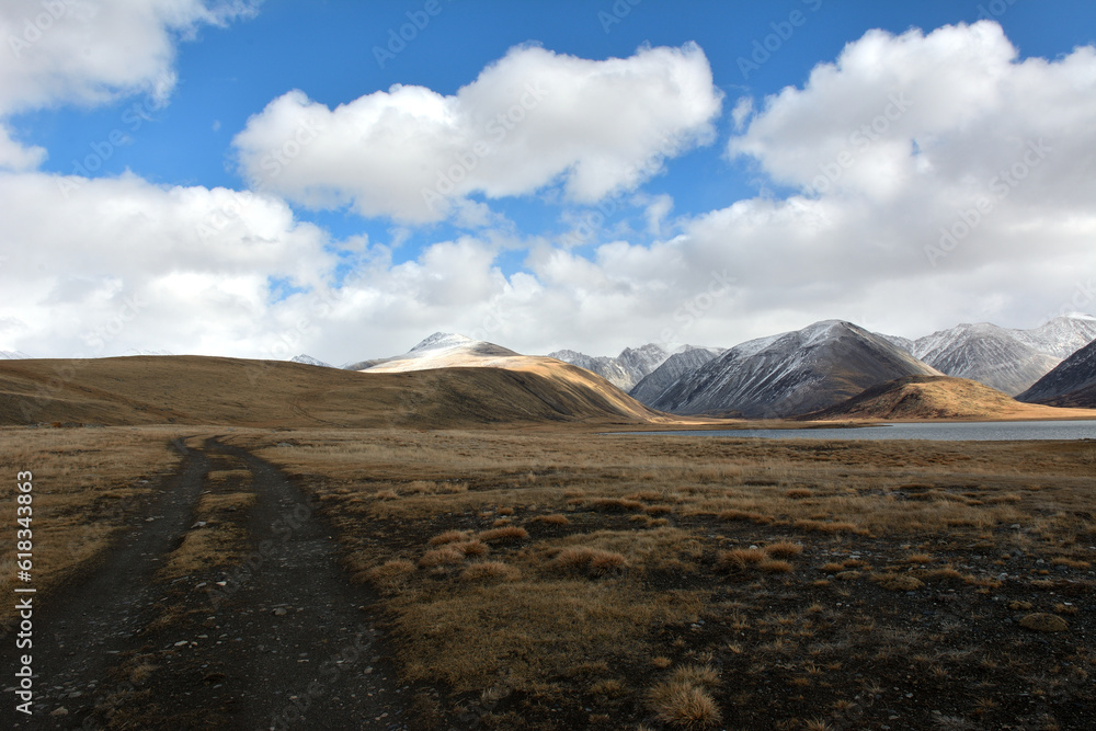 A winding field road rises from the lowland to the hills on the shore of a large beautiful lake lying in the steppe at the foot of high snow-capped mountains. A winding field road rises from the lowland to the hills on the shore of a large beautiful lake lying in the steppe at the foot of high snow-capped mountains.