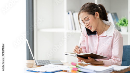 Photo of adorable young lady talking on mobile phone and writing note, working at home office.