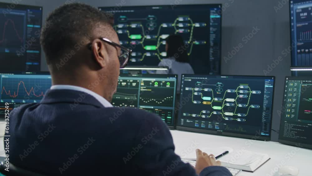 Software programmer sits in front of computer with blockchain network and data server in monitoring office. Team of IT technical specialists work with live analysis charts on big digital screens. Software programmer sits in front of computer with blockchain network and data server in monitoring office. Team of IT technical specialists work with live analysis charts on big digital screens.