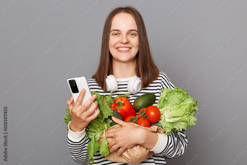 Nutritious grocery purchases for a balanced diet. Online ordering products. Happy woman using mobile phone embracing fresh vegetables returns from supermarket wearing striped shirt. Nutritious grocery purchases for a balanced diet. Online ordering products. Happy woman using mobile phone embracing fresh vegetables returns from supermarket wearing striped shirt.