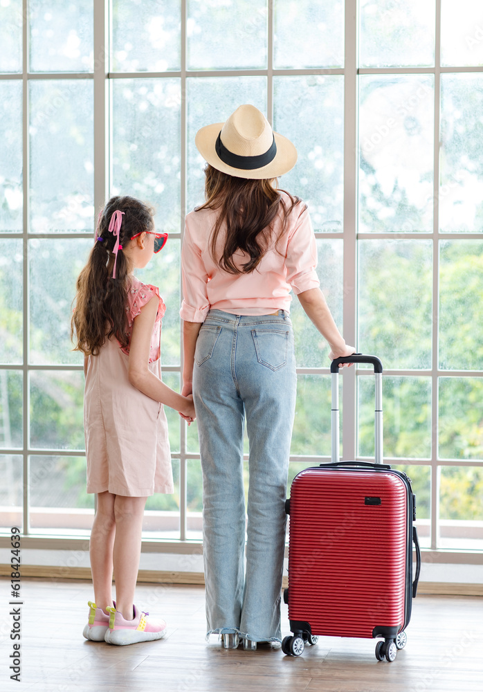 Asian unrecognizable unknown family mom and daughter wearing sunglasses and hat standing with red trolley luggage waiting for departure time in airport hallway for traveling vacation weekend trip