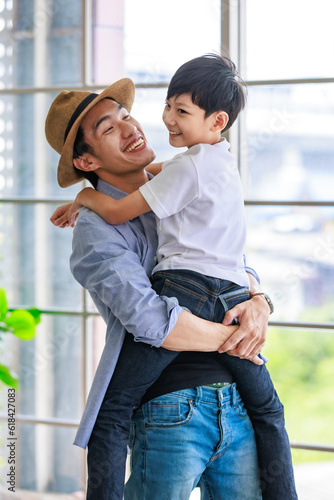 Asian happy cheerful joyful family young father wearing hat standing smiling bonding holding hugging cuddling lifting little preschooler boy son playing funny together in apartment home living room © Bangkok Click Studio