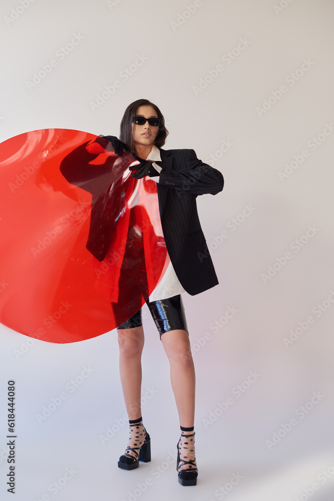 Fototapeta premium pretty asian model in stylish look and sunglasses posing holding red round shaped glass, grey background, blazer and latex shorts, youthful and modern woman, fashion statement, studio photography