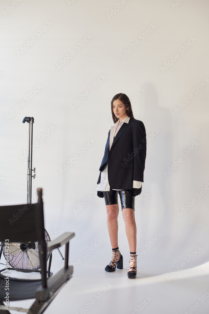 edgy fashion, studio photography, young asian woman in stylish look posing on grey background, blazer, white shirt and latex shorts, standing near electric fan, personal style, full length