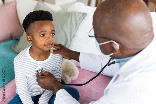 Senior african american male doctor in face mask examining boy patient using stethoscope photo