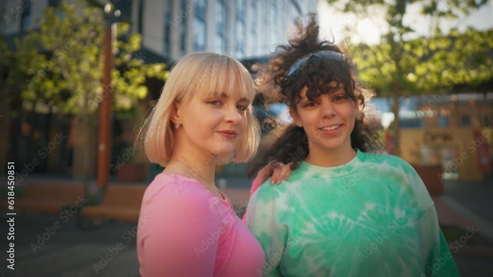 Two Stylish Female Teenagers Are Looking At Camera And Smiling On The City Street. Young Fashionable Women Relaxing Outside In Downtown District During Sunny Summer Day. Youth Culture Concept. Two Stylish Female Teenagers Are Looking At Camera And Smiling On The City Street. Young Fashionable Women Relaxing Outside In Downtown District During Sunny Summer Day. Youth Culture Concept.