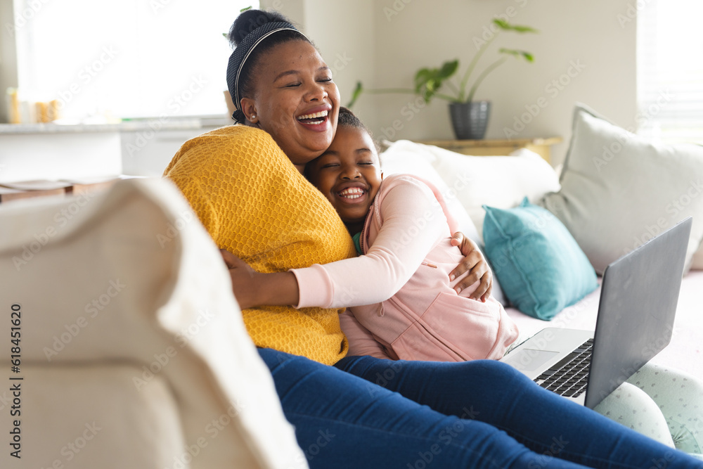Happy african american mother and daughter sitting on sofa, embracing and using laptop at home Happy african american mother and daughter sitting on sofa, embracing and using laptop at home