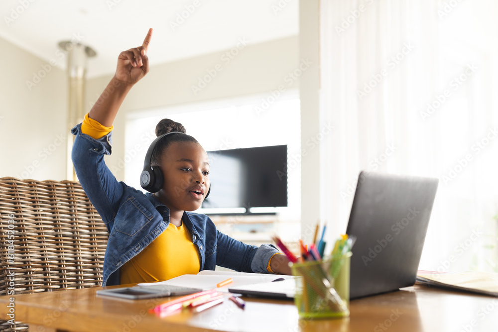 African american girl sitting at table using laptop for online lesson and raising hand