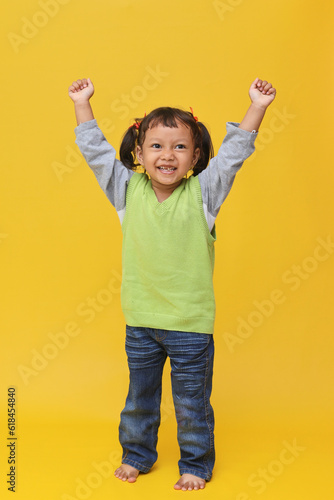 Full body happy asian little girl isolated on vivid yellow background raising her fists, hands up