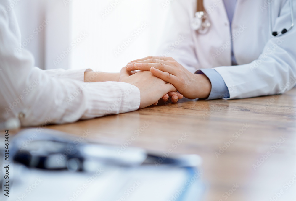 Obraz premium Doctor and patient sitting at the wooden table in clinic. Female physician's hands reassuring woman. Medicine concept