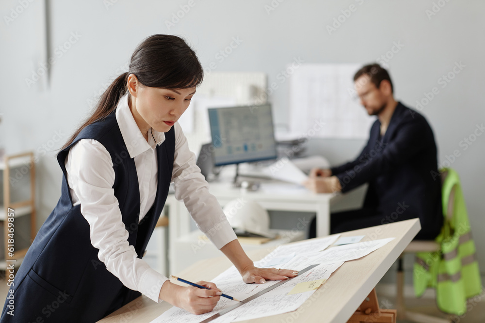 Fototapeta premium Side view portrait of young woman as engineer drawing blueprints and plans at table in office, copy space