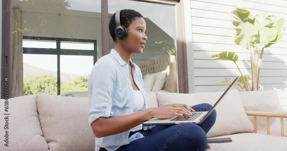 African american woman sitting on sofa on terrace wearing headphones, using laptop, slow motion African american woman sitting on sofa on terrace wearing headphones, using laptop, slow motion