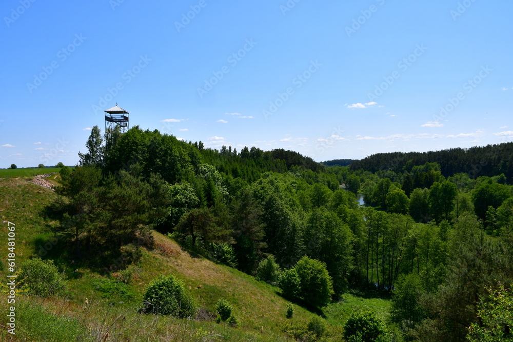 A close up on a tall observational or sentry tower made out of metal and wood standing in the middle of a vast field full of crops and a lush forest or moor spotted on a sunny summer day in Poland