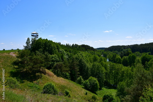 A close up on a tall observational or sentry tower made out of metal and wood standing in the middle of a vast field full of crops and a lush forest or moor spotted on a sunny summer day in Poland
