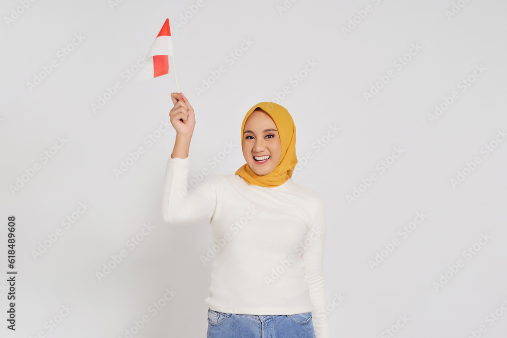 Excited young Asian Muslim woman in hijab celebrate Indonesian independence day on 17 August and holding the Indonesian flag isolated over white background Excited young Asian Muslim woman in hijab celebrate Indonesian independence day on 17 August and holding the Indonesian flag isolated over white background