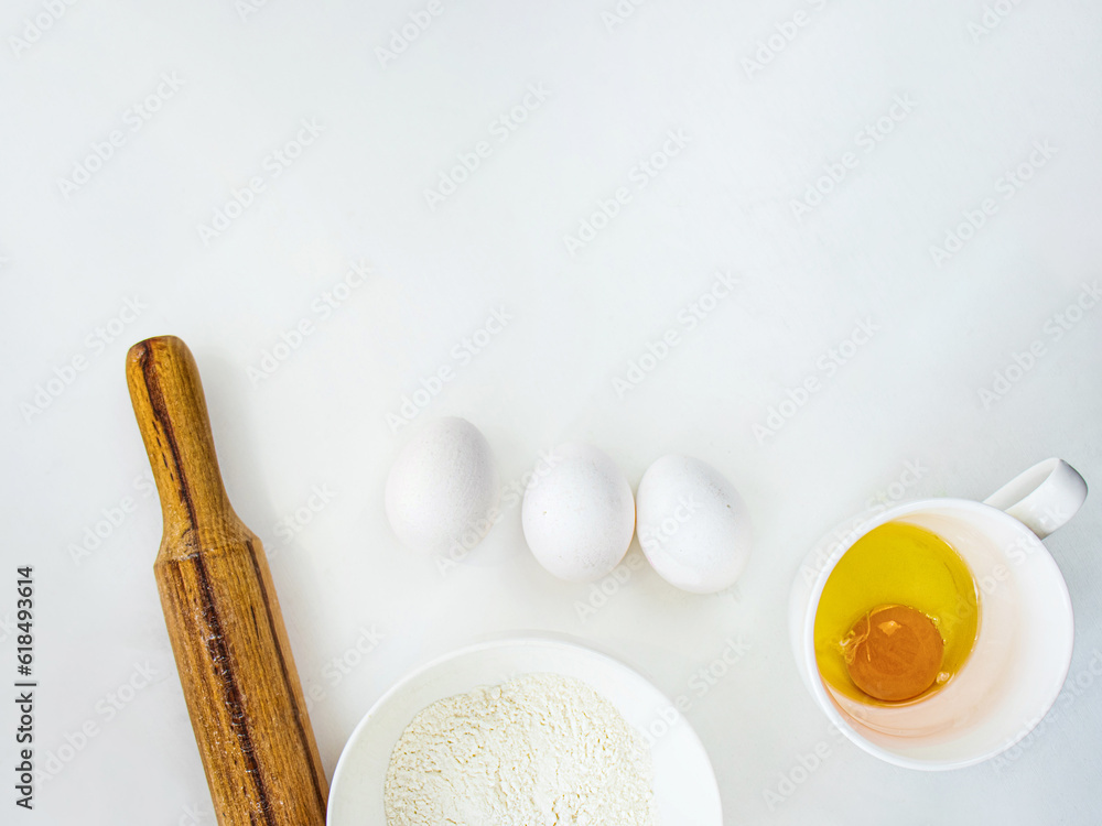 Fototapeta premium chicken eggs, a wooden rolling pin, flour in a bowl and a glass with a raw egg on the table, dough recipe, in the kitchen at home, on a white background, copy space, dough preparation