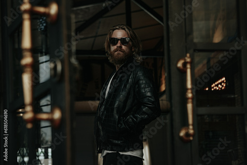 Brutal caucasian man with long beard hipster in black leather jacket stay next to glass door