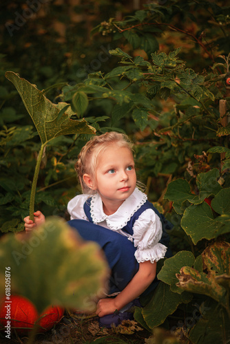 A little blonde girl in a blue rustic dress is sitting under a pumpkin leaf