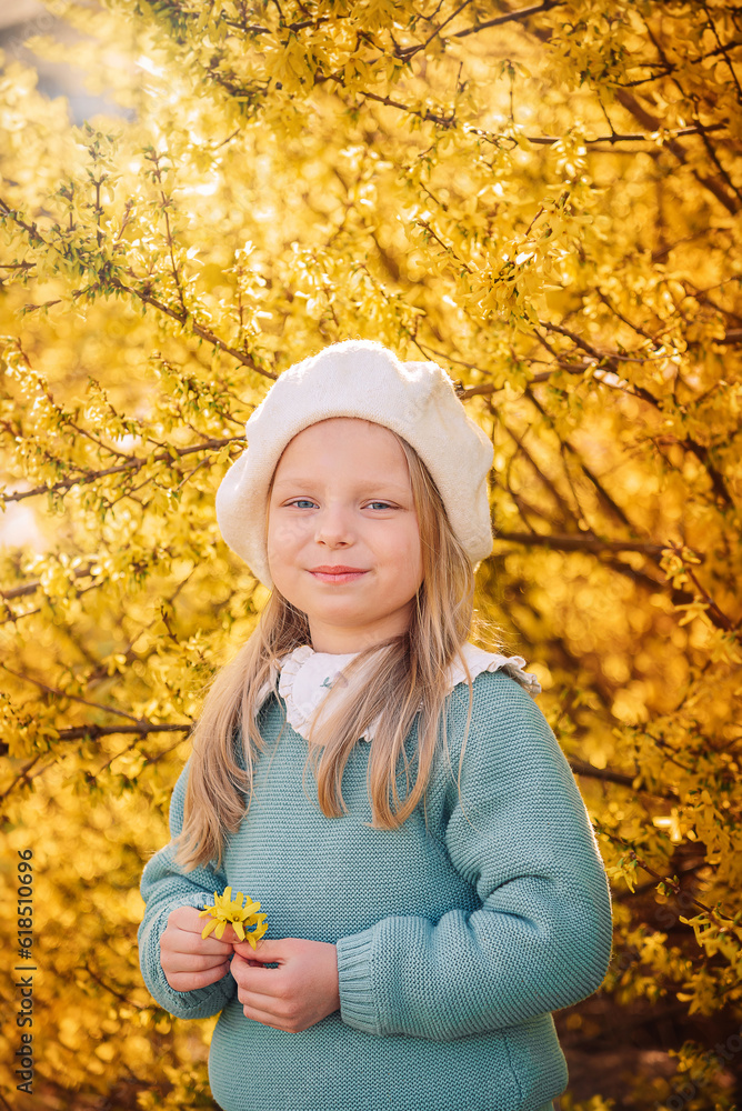 A little blonde girl in a white beret and a turquoise sweater stands against the background of a blooming yellow forsythia A little blonde girl in a white beret and a turquoise sweater stands against the background of a blooming yellow forsythia