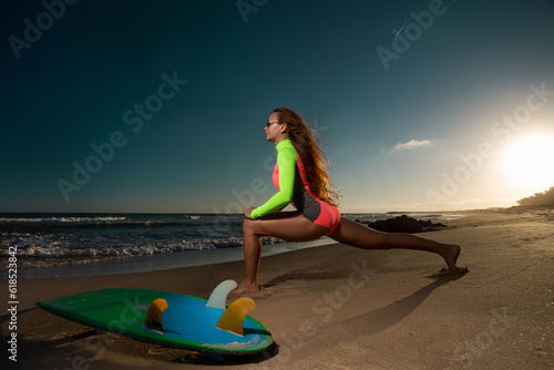 Young woman in swimsuit exercising on the beach with surfboard in sunset