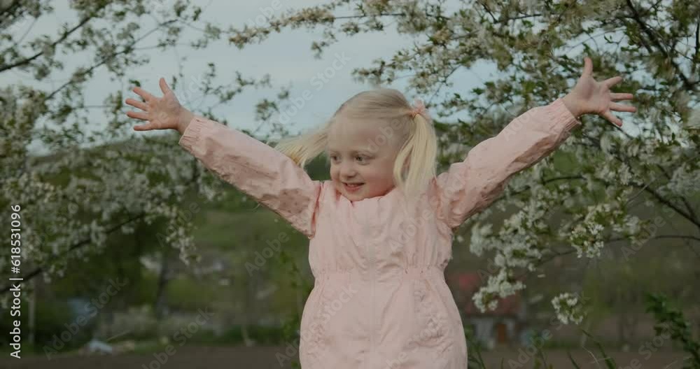 Little blond girl with two ponytails raises her hands and smiles. Flower petals fall from trees. Happy springtime Little blond girl with two ponytails raises her hands and smiles. Flower petals fall from trees. Happy springtime