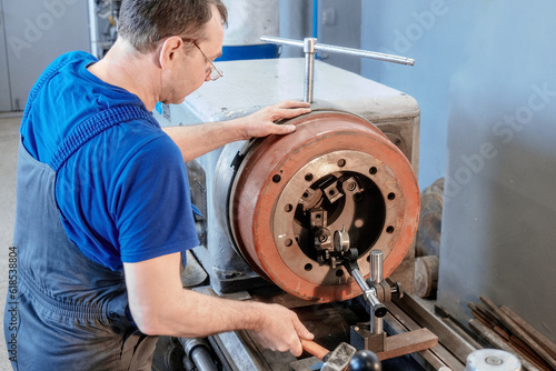 An elderly Caucasian metal turner works in a workshop behind a lathe. Authentic workflow. A real defense industry worker works in a factory.
