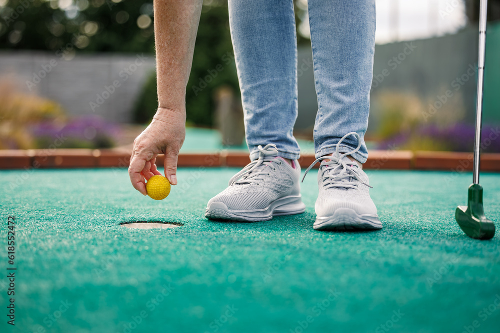 Woman plays mini golf and taking ball out from the hole after successful putting - obrazy, fototapety, plakaty 