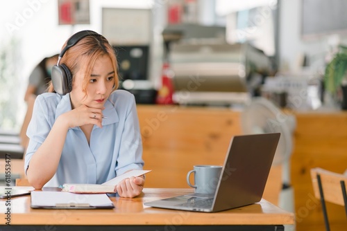 A smiling student girl wears wireless headphones, studying online in a cafe. Learning language, listening to lectures, watching webinars, and taking notes. Represents distance education.
