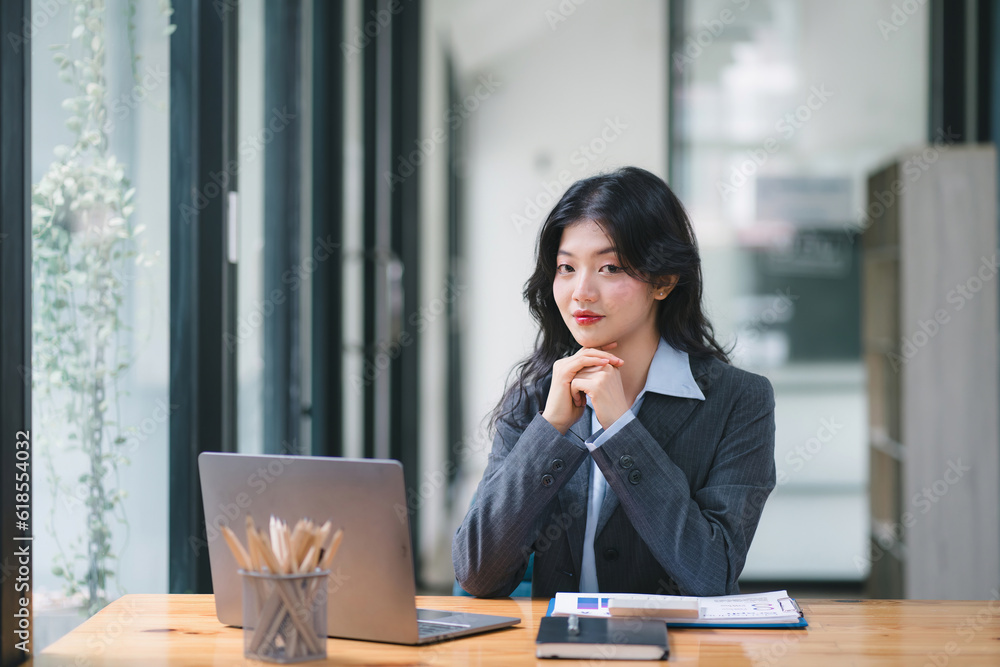 Portrait of a successful and lovely millennial Asian businesswoman, secretary, sales manager, or business consultant, sitting at her workplace with a laptop, smiling, while dressed casually. Portrait of a successful and lovely millennial Asian businesswoman, secretary, sales manager, or business consultant, sitting at her workplace with a laptop, smiling, while dressed casually.