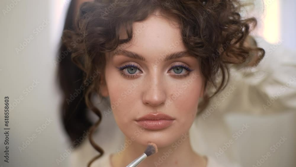 Closeup portrait of young sensual woman looking at camera during professional makeup in modern beauty studio. Hairdresser doing stylish hairdo during four-handed makeup and styling, slow motion. Closeup portrait of young sensual woman looking at camera during professional makeup in modern beauty studio. Hairdresser doing stylish hairdo during four-handed makeup and styling, slow motion.