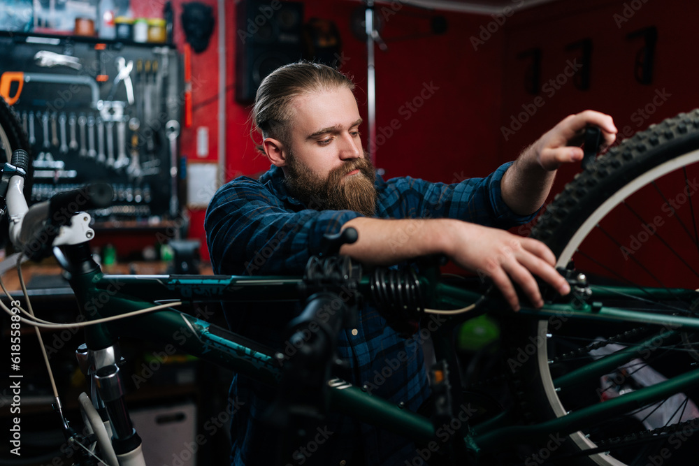 Portrait of bearded cycling mechanic male repairing and fixing mountain bicycle standing on bike rack working in bicycle repair shop with dark interior. Concept of maintenance of bicycle transport. Portrait of bearded cycling mechanic male repairing and fixing mountain bicycle standing on bike rack working in bicycle repair shop with dark interior. Concept of maintenance of bicycle transport.