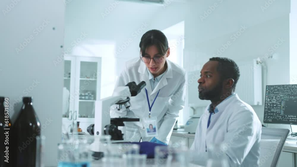 In the laboratory two scientists in uniform looking under a microscope talking discuss wonder. African American man and a Caucasian woman. Research, biochemistry, pharmaceutical medicine In the laboratory two scientists in uniform looking under a microscope talking discuss wonder. African American man and a Caucasian woman. Research, biochemistry, pharmaceutical medicine