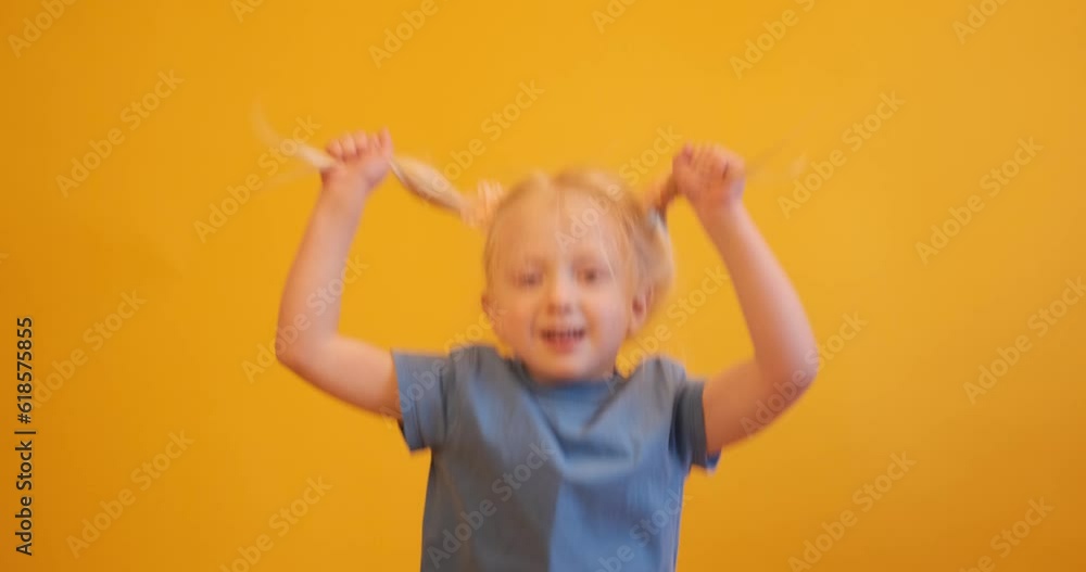 Little fair-haired girl with two ponytails holding herself by the hair and jumping on yellow background. Happy childhood Little fair-haired girl with two ponytails holding herself by the hair and jumping on yellow background. Happy childhood