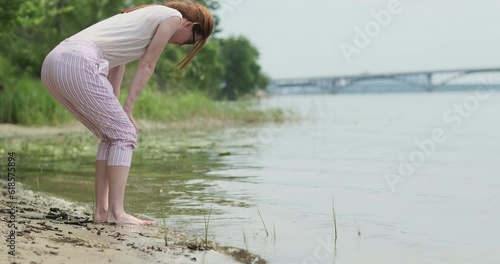 Young woman examines living creatures near river bank. Girl bent over water on shore, summer, river, bridge, green trees.