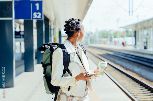 Woman with afro hair using smartphone while wait for her train on the railway station platform. Tourism and travel in the summer. Vacations for the student. Work and travel.
