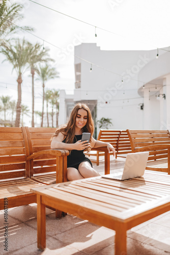 A woman remotely works on the cafe terrace with a laptop and phone in a country where it is always summer. A girl with an online profession works from anywhere in the world, travels in a warm climate